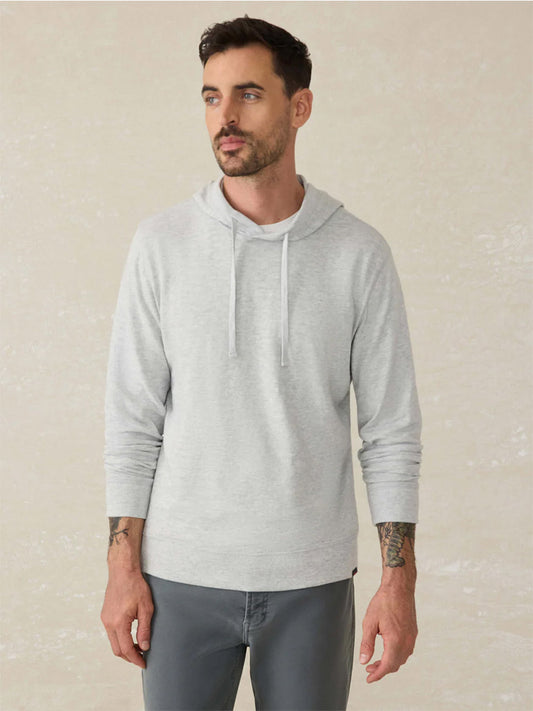 A man wears the Faherty Brand Sunwashed Midweight Organic Hoodie in Light Grey Heather with drawstrings and gray pants, standing against a plain light background.