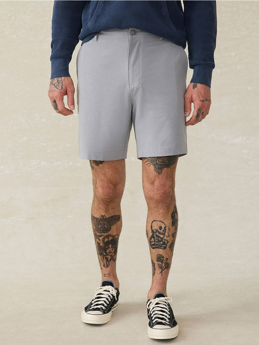 A person wearing a navy sweatshirt, Faherty Brand All Day Short in Ice Grey, black-and-white sneakers, and showing tattoos on their hands and legs stands against a neutral background.
