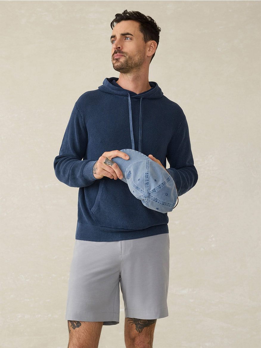 A man in a navy hoodie and Faherty Brand All Day Short in Ice Grey, known for their water-repellent design, holds a light blue baseball cap while standing against a plain light-colored background.