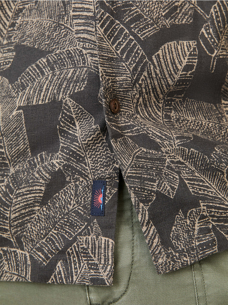 Close-up of the Faherty Brand Short-Sleeve Breeze Camp Shirt in Graphite Rainforest, featuring a dark patterned cotton blend with beige leaf designs, camp collar, button detail, fabric tag, and olive green pants beneath the hem.