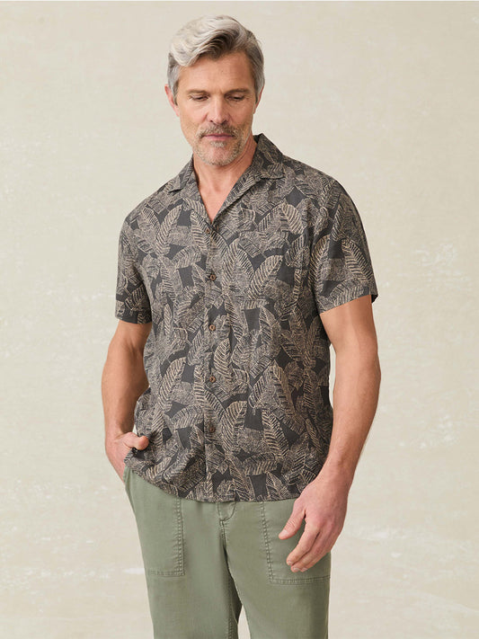 A man with gray hair wears the Faherty Brand Short-Sleeve Breeze Camp Shirt in Graphite Rainforest and olive green pants, standing with one hand in his pocket against a light background for a subtle throwback style.