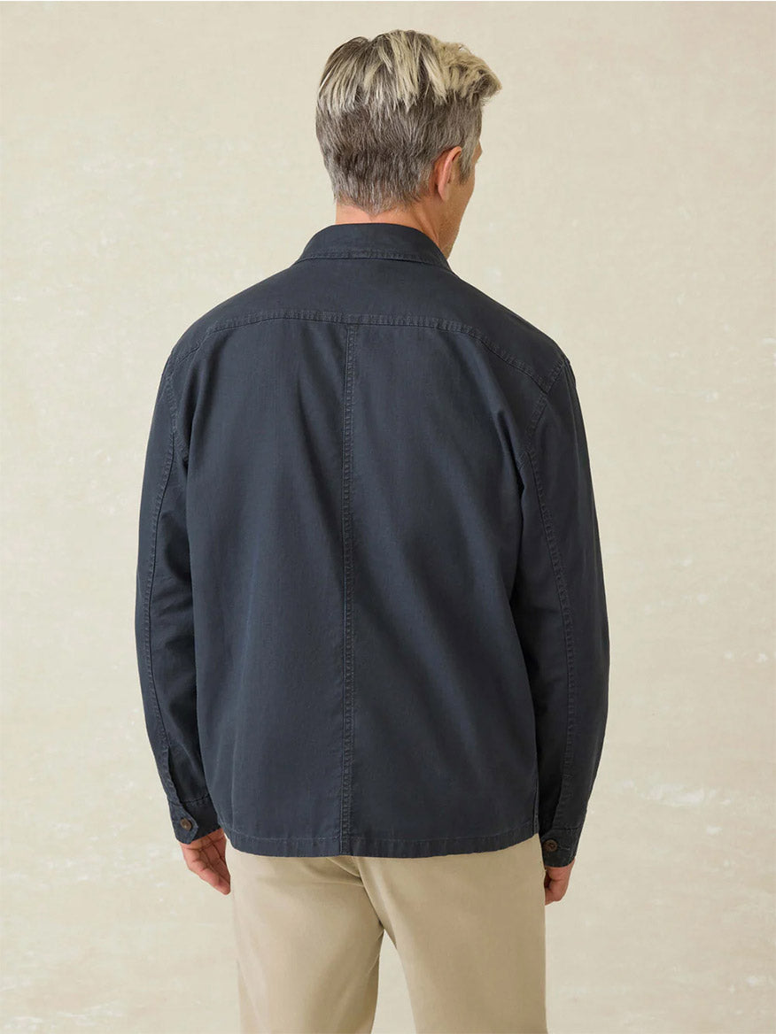 A man with short, light hair is seen from behind wearing the Faherty Brand Commander Shirt Jacket in Washed Charcoal, made from breathable organic cotton-linen for durability, along with beige pants, standing against a neutral background.