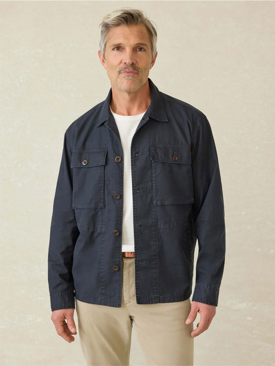 A man with gray hair and a short beard wears the Faherty Brand Commander Shirt Jacket in Washed Charcoal over a white t-shirt and beige pants, standing against a plain light background.