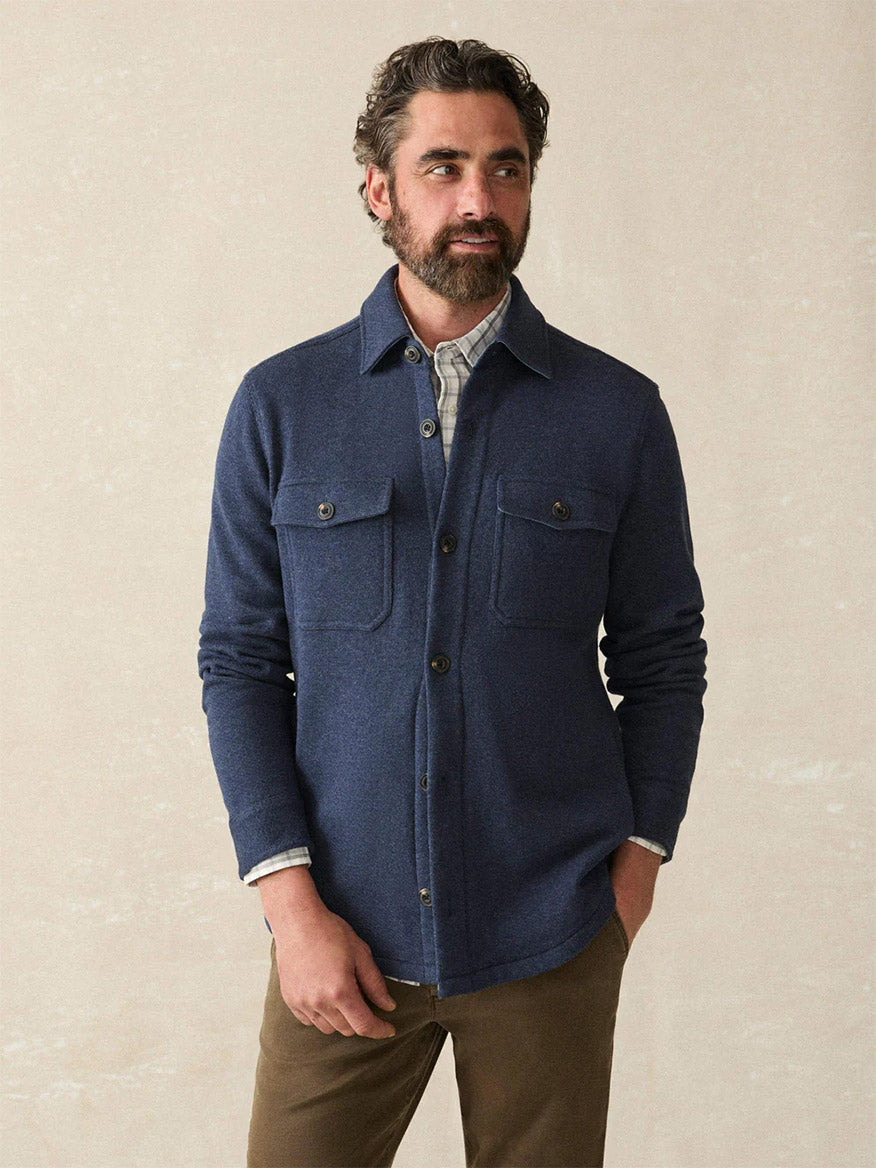 A man with short curly hair and a beard wears the Faherty Brand Inlet Knit CPO in Navy Night Melange over a plaid shirt with brown pants, standing against a plain light background.
