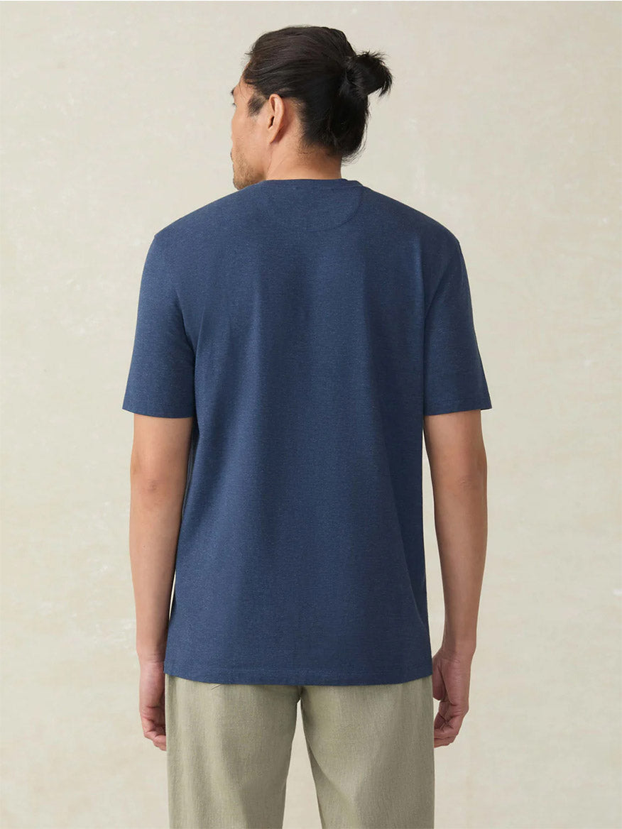 A man with dark hair in a bun stands with his back to the camera, wearing the Faherty Brand Movement Short-Sleeve T-Shirt in Great Falls Heather and light-colored pants against a plain background.