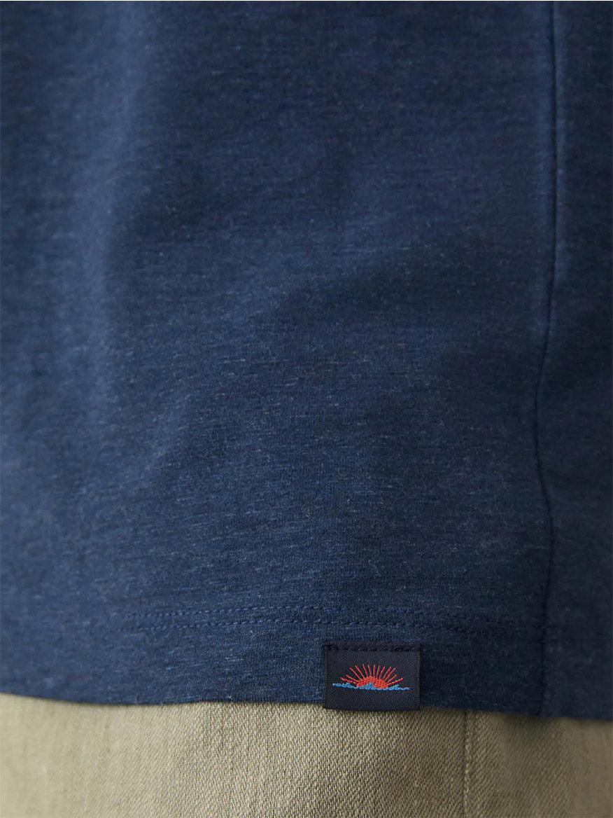 Close-up of the hem of the Faherty Brand Movement Short-Sleeve T-Shirt in Great Falls Heather, featuring a small rectangular tag with a red and blue sunrise design, worn above light-colored pants.