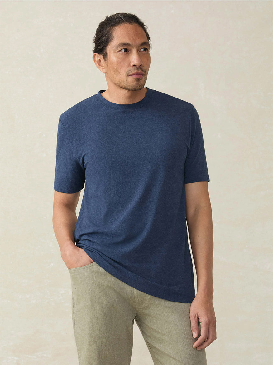 A man with long dark hair in a ponytail wears the Faherty Brand Movement Short-Sleeve T-Shirt in Great Falls Heather and light beige pants, standing with one hand in his pocket against a neutral background.