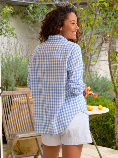 A woman in a Frank & Eileen Eileen Relaxed Button-Up Shirt in Blue Check and white shorts smiles over her shoulder in a garden patio setting.
