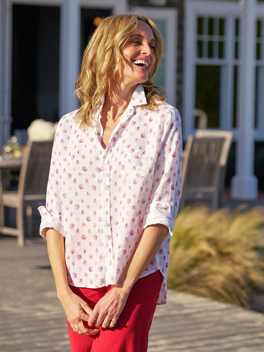 A woman stands outdoors, smiling in the Frank & Eileen Eileen Relaxed Button-Up Shirt in Red & Blue Flower and red pants, embodying casual California effortlessness. Wooden chairs and a table are visible in the background.