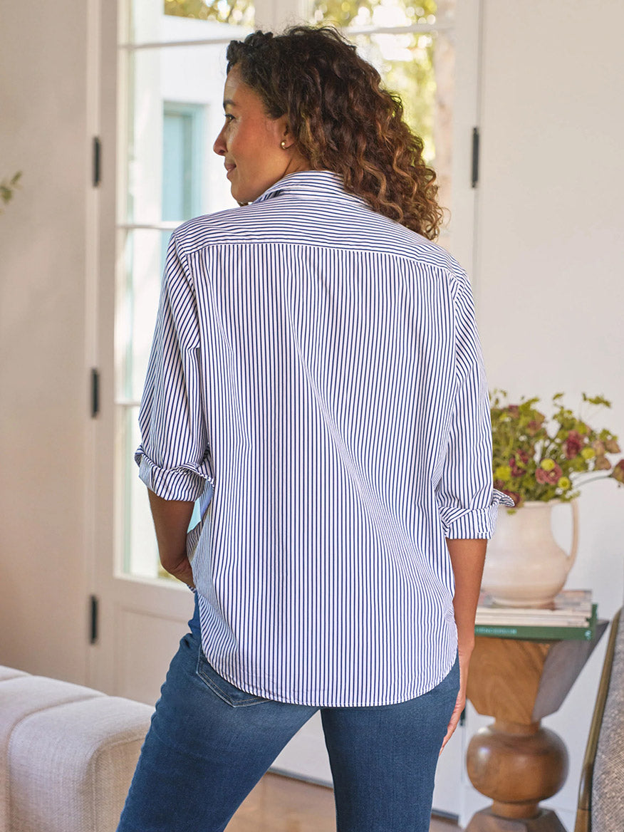 A woman stands indoors with her back to the camera, wearing the Frank & Eileen Eileen Relaxed Button-Up Shirt in White & Navy Stripe and jeans. A vase of flowers beside her adds a touch of effortless California style.