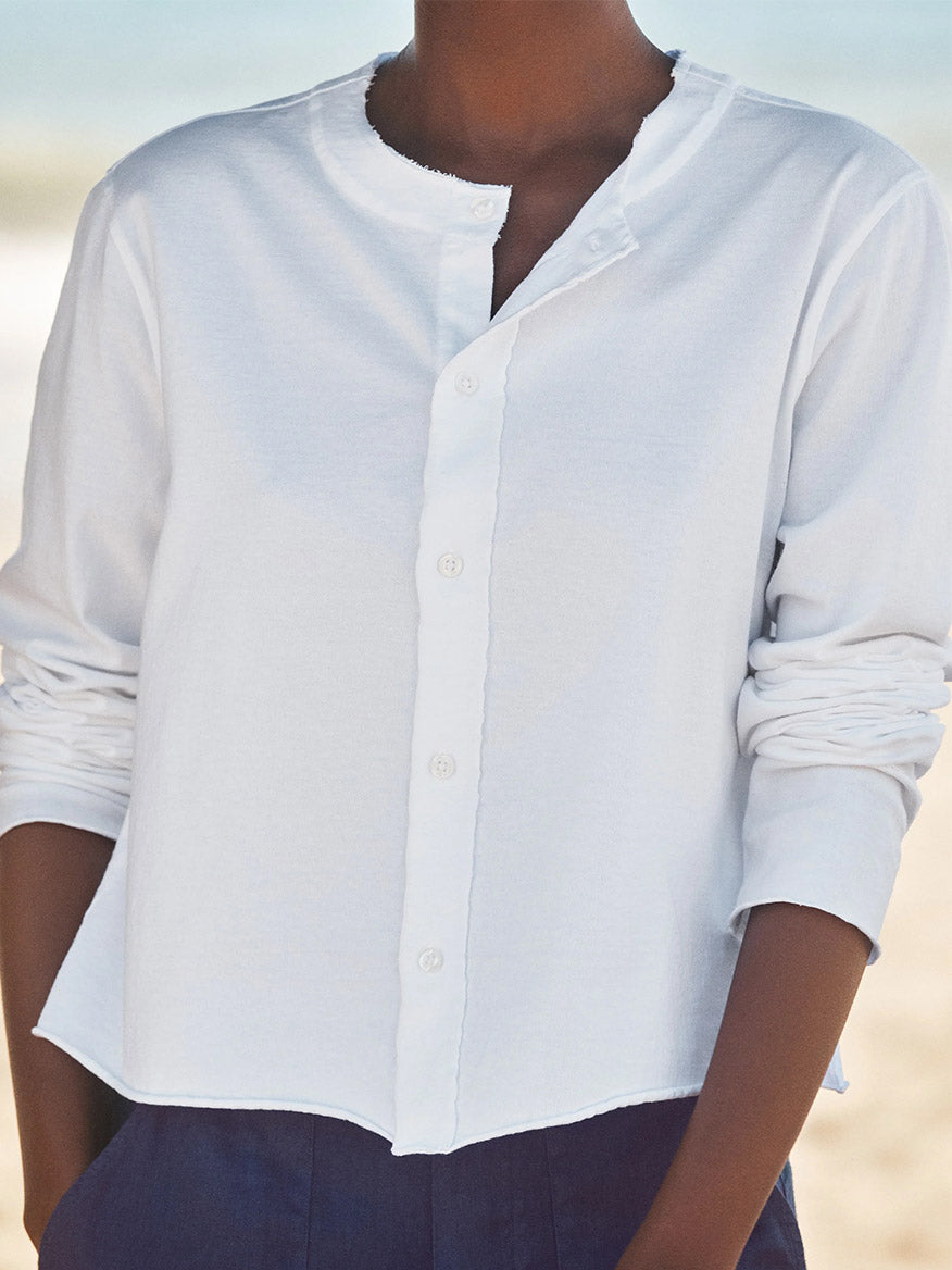 A person outdoors with a blurred background wears the Frank & Eileen Jackie T-Shirt Cardigan in White Vintage Jersey draped over their shoulders, paired with a long-sleeve white button-up shirt with rolled sleeves.