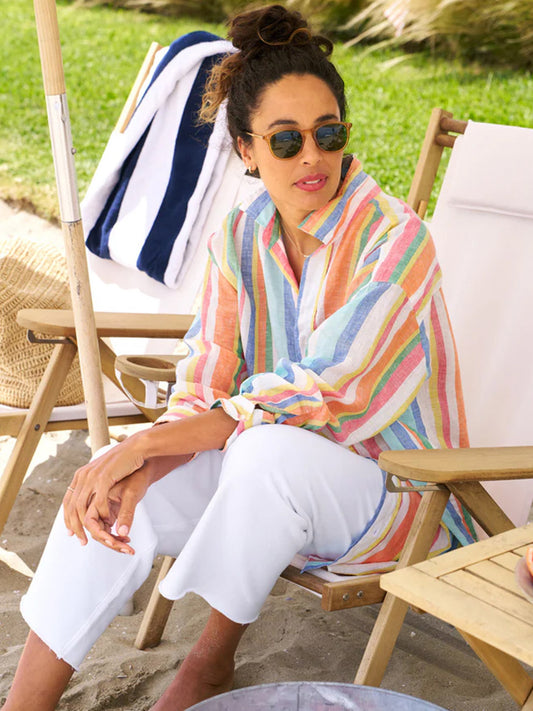 A woman sits on a beach chair wearing a Frank & Eileen Mackenzie One-Size Button-Up Shirt in Multi Stripe Linen and white pants, sunglasses on, with a relaxed expression.
