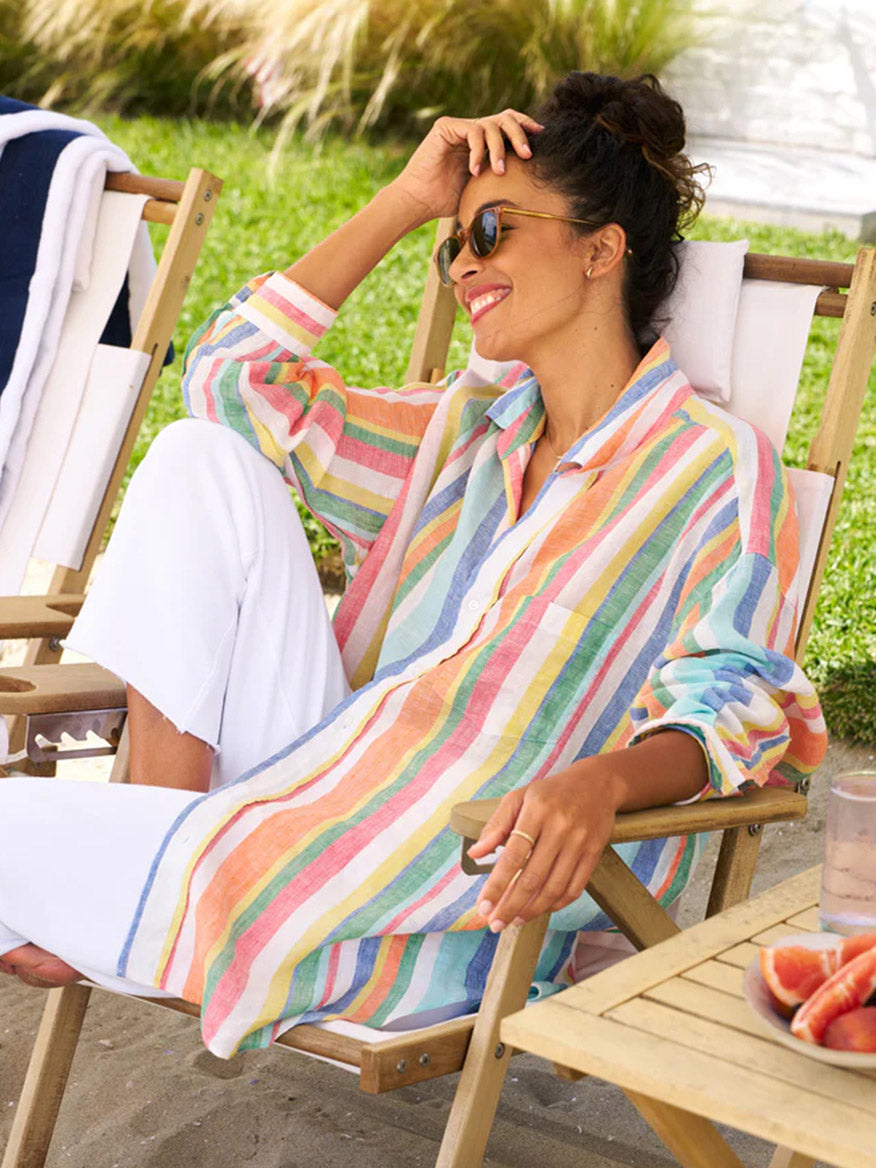 A woman relaxing in a deck chair by the beach, wearing a Frank & Eileen Mackenzie One-Size Button-Up Shirt in Multi Stripe Linen and white pants, smiling and shielding her eyes from the sun.