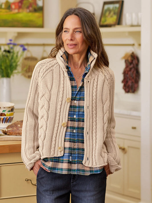 Woman stands in a kitchen wearing the Frank & Eileen Mini Cotswolds Cropped Cable Knit Cardigan in Sand over a blue plaid shirt and dark jeans, hands in her pockets, looking to the side.
