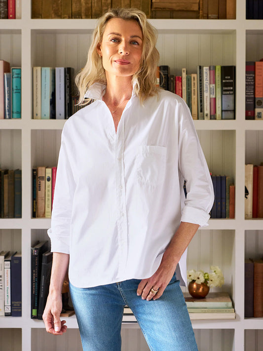 A woman with blonde hair wears the Frank & Eileen Oona Continuous Sleeve Button-Up Shirt in White and blue jeans as she stands before a bookshelf filled with books.