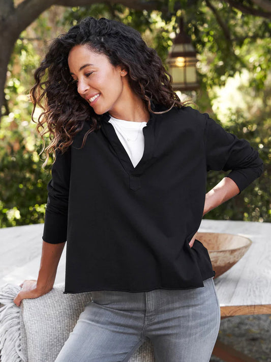 A woman with curly hair, smiling, wearing a Frank & Eileen Patrick Popover Henley in Black and grey pants, stands outdoors, leaning on a cushioned bench.