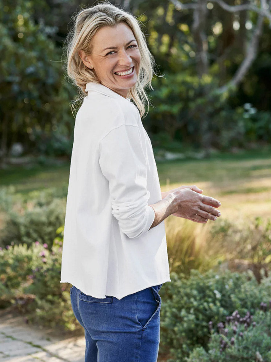 A smiling woman with blond hair, wearing a Frank & Eileen Patrick Popover Henley in White Heritage Jersey and blue jeans, standing outdoors.
