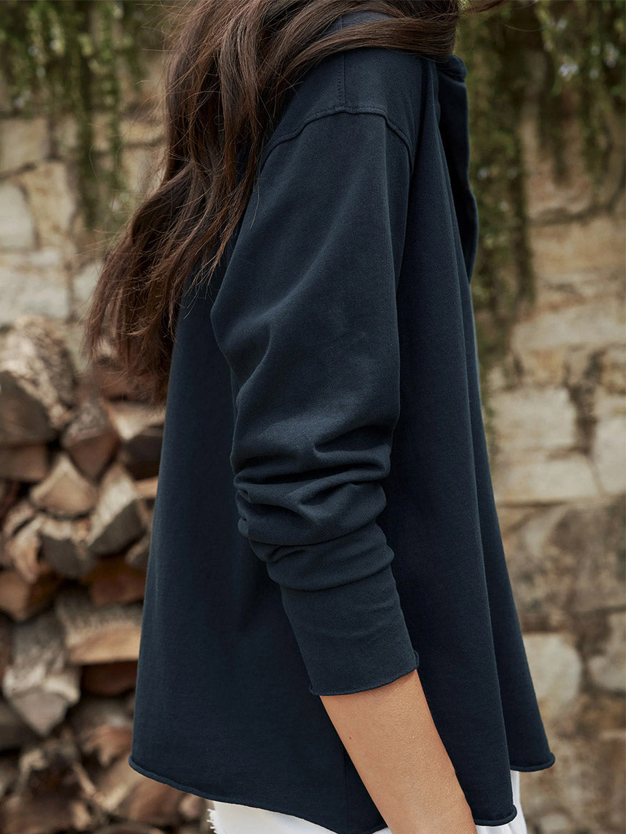 Close-up of a woman in a Frank & Eileen Patrick Popover Henley in British Royal Navy, focusing on the sleeve and back. She is facing a stone wall layered with chopped wood.