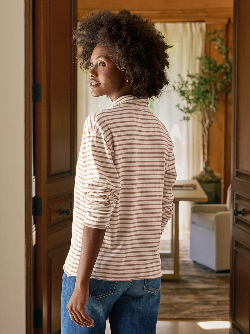 A person with curly hair, wearing the Frank & Eileen Patrick Popover Henley in Vintage White & Camel Stripe Heritage Jersey and jeans, stands in a doorway looking back into a sunlit room with a plant.