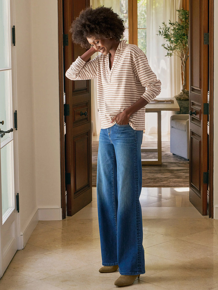 Indoors, a person smiles while touching their head, wearing the Frank & Eileen Patrick Popover Henley in Vintage White & Camel Stripe Heritage Jersey with wide-leg blue jeans and tan shoes.