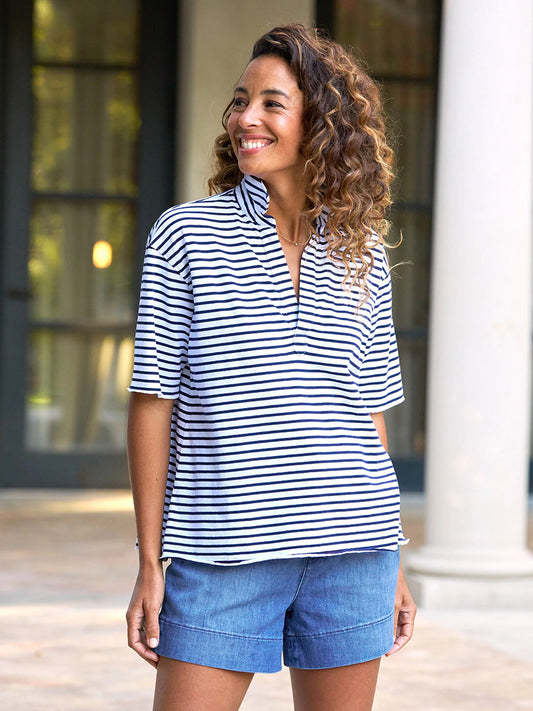 Patty, with curly hair, smiles outdoors in front of a building wearing the Frank & Eileen Patty Short-Sleeve Popover Henley in Navy French Stripe and blue denim shorts.
