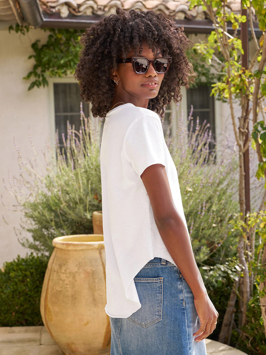 A woman with curly hair and sunglasses wears the Frank & Eileen Theo Perfect Tee in White Heritage Jersey and blue jeans, standing outdoors by plants and a large ceramic pot.