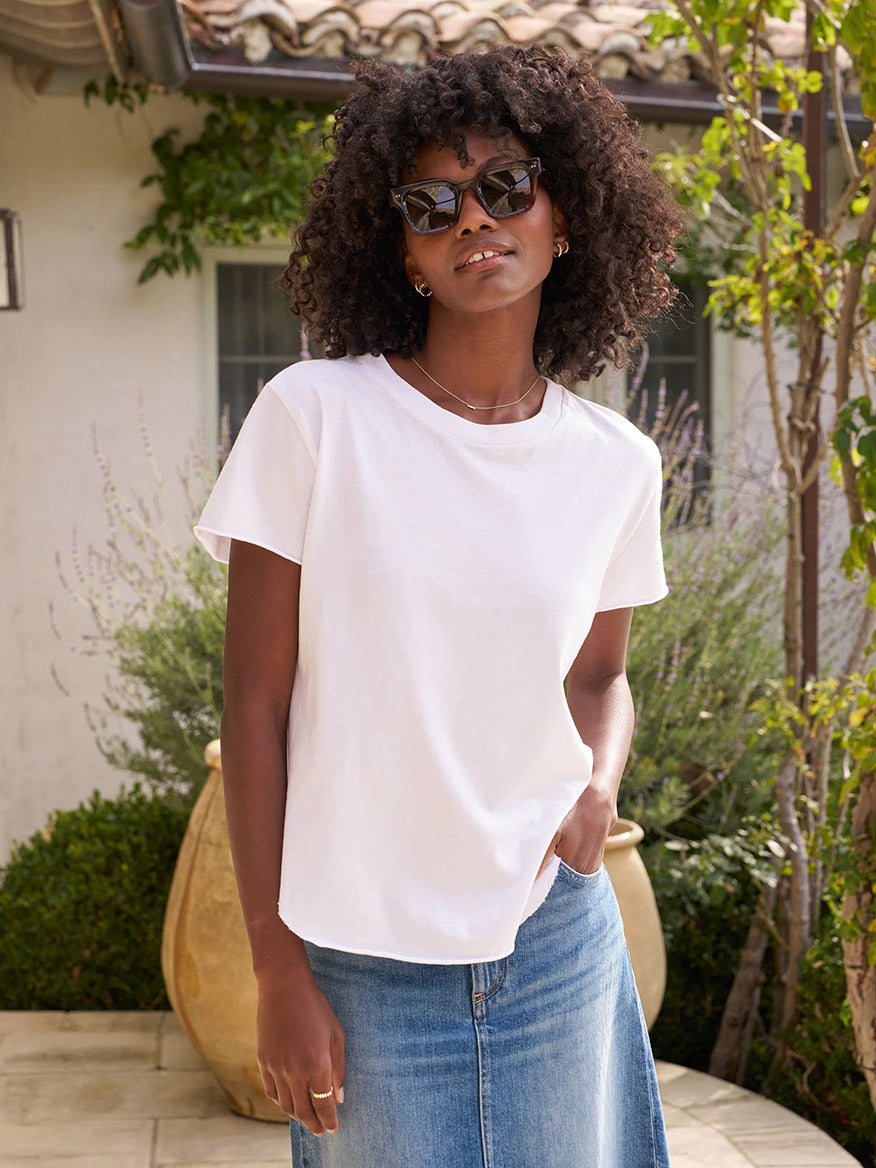 A woman in sunglasses and a denim skirt stands outdoors, wearing the Frank & Eileen Theo Perfect Tee in White Heritage Jersey, with one hand in her pocket.