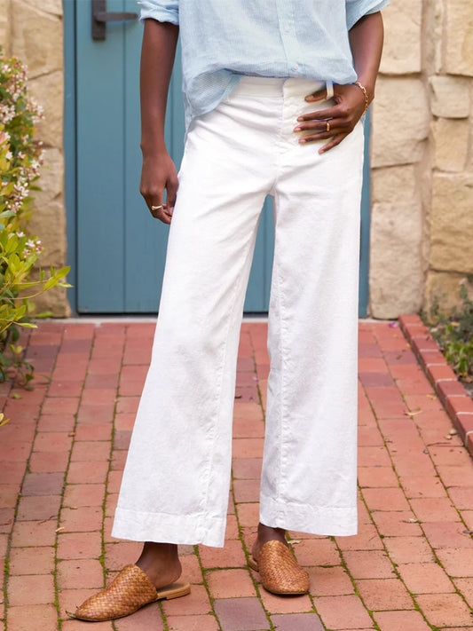 A person stands in a courtyard wearing Frank & Eileen Wexford Wide-Leg Linen pants in White, a light blue shirt, and woven brown loafers, holding a smartphone. Visible from waist to toes.