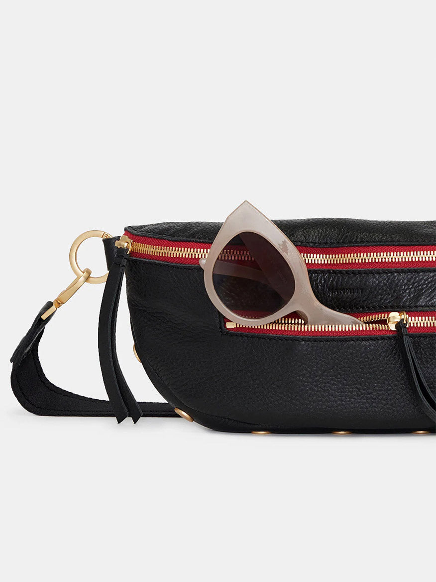 A Hammitt Los Angeles Charles Crossbody Medium in black with brushed gold accents and a red zippered pocket, featuring a chic pair of sunglasses with beige frames.