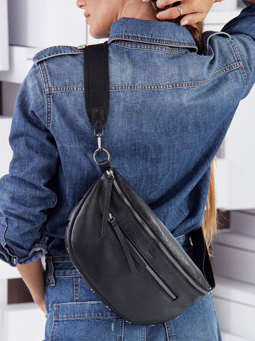 A woman in a denim jacket carries a Hammitt Los Angeles Charles Crossbody Large in Black over her shoulder, posing with her hand in her hair, viewed from behind.