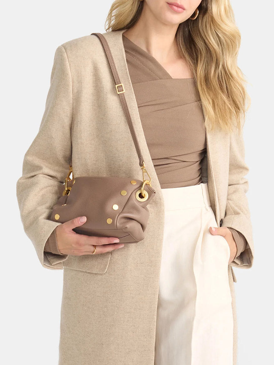A person in a tan coat holds the Hammitt Los Angeles Daniel Crossbody Clutch Small in Sculpted Taupe, a brown handbag with gold studs. They wear a beige top and white pants against a plain background.