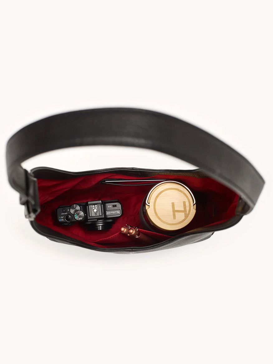 The Hammitt Los Angeles Tim Hobo in Black, featuring a red interior, holds a camera, a round container marked “H,” and beaded earphones.