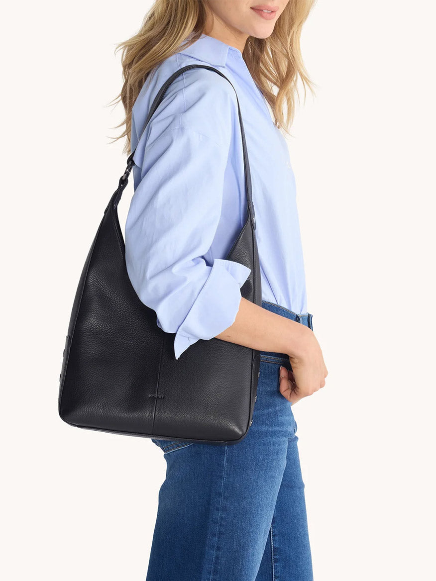 A woman in a light blue shirt and jeans carries the Hammitt Los Angeles Tim Hobo in Black on her right shoulder.