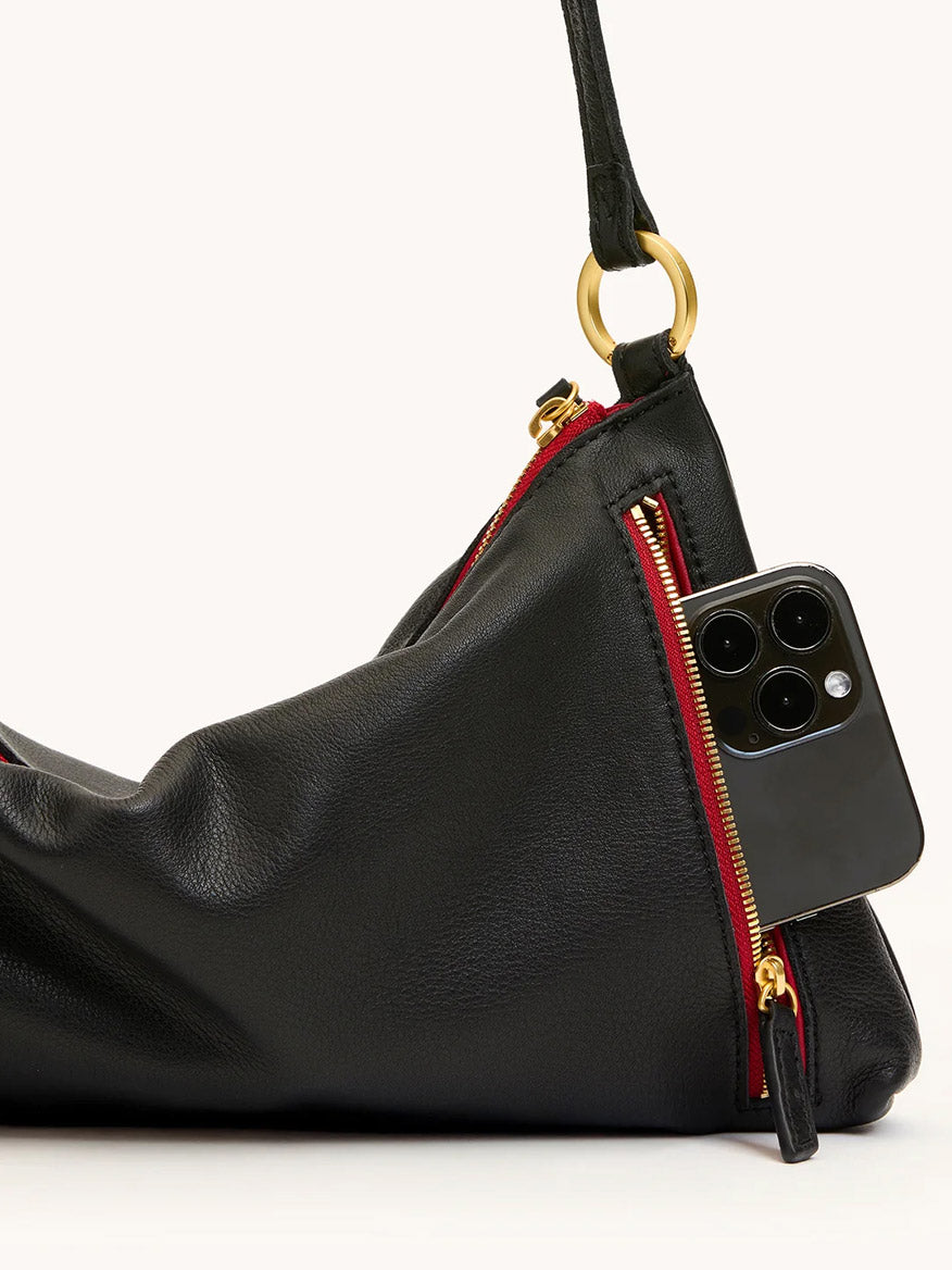 The Hammitt Los Angeles Kyle Medium Shoulder Bag in Black/Brushed Gold/Red Zip is styled as a crossbody and shoulder bag, with a smartphone shown partially tucked into one of its zippered pockets.