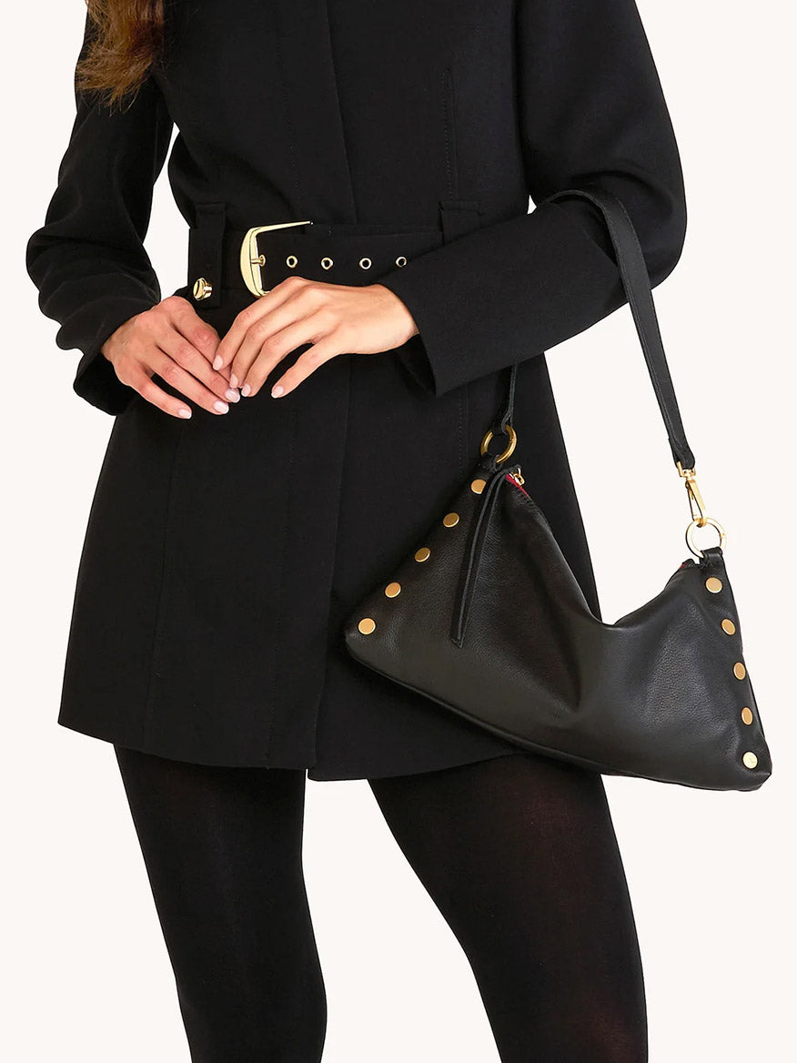 Woman wearing a black belted coat holds the Hammitt Los Angeles Kyle Medium Shoulder Bag in Black/Brushed Gold/Red Zip, a versatile bag with gold details that can be worn as a crossbody or shoulder bag.