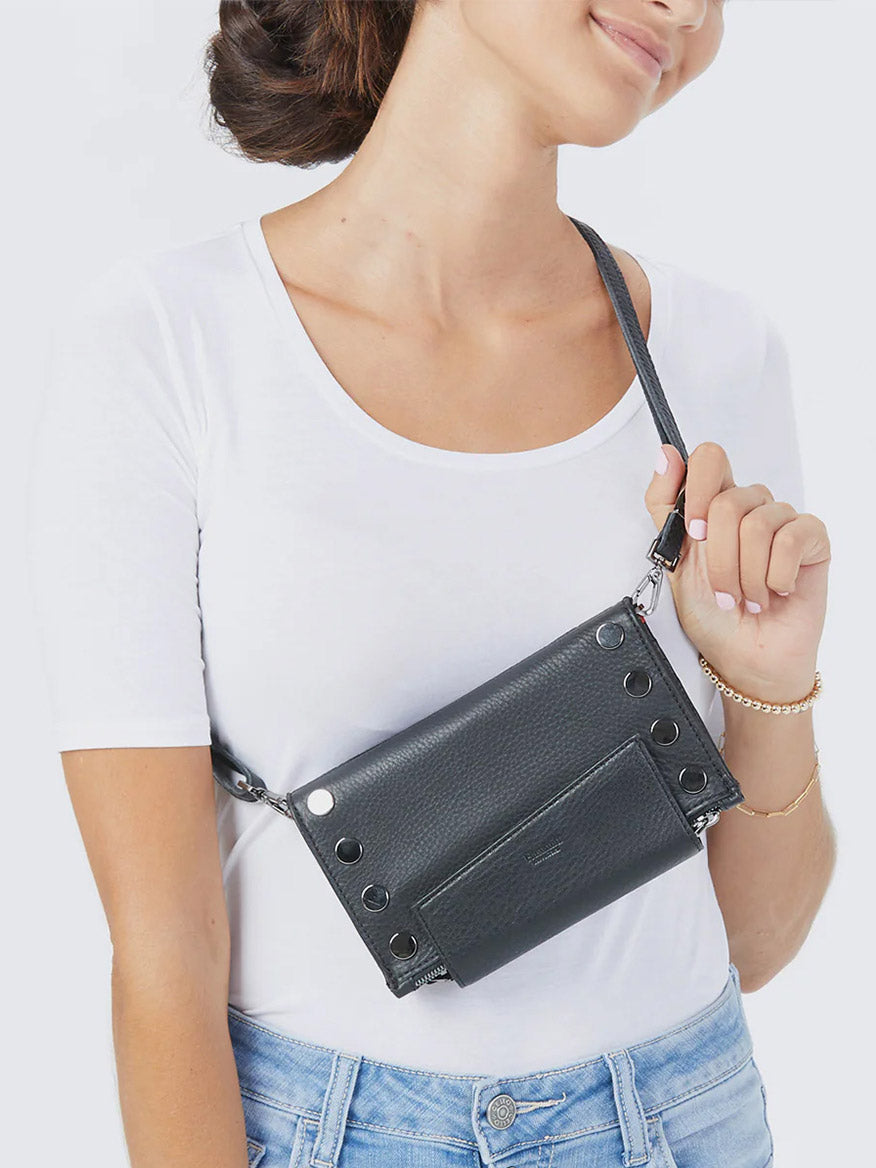 A woman in a white t-shirt and jeans carries the Hammitt Los Angeles Levy Clutch in Black/Gunmetal, featuring grommet details and a removable adjustable strap over her shoulder.