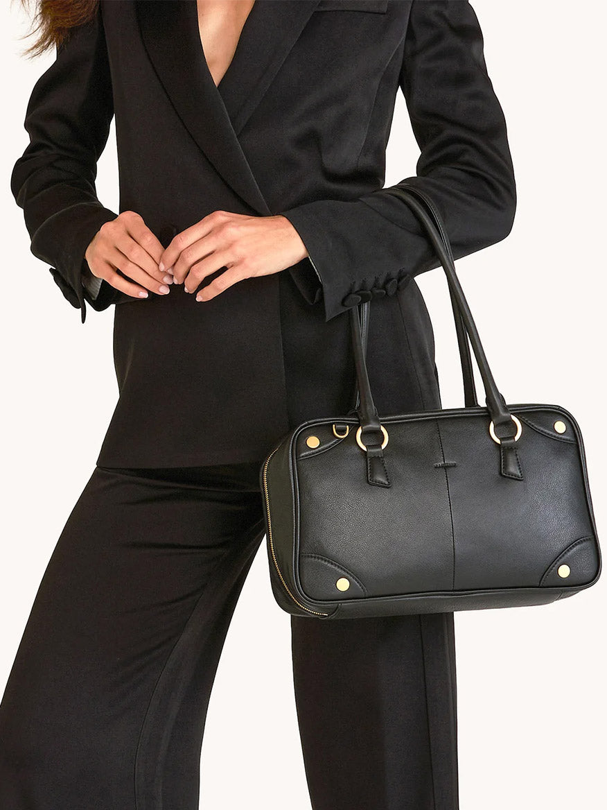 A woman in a black suit holds the Hammitt Los Angeles Meredith Medium in Black/Brushed Gold, featuring gold rivet details, while standing against a plain light background.