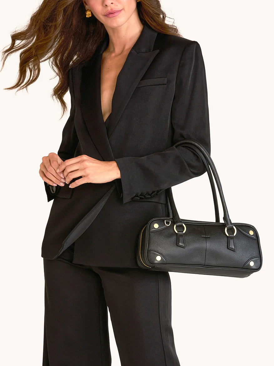 A person in a black suit carries the Hammitt Los Angeles Meredith Small in Black/Brushed Gold handbag over their arm.