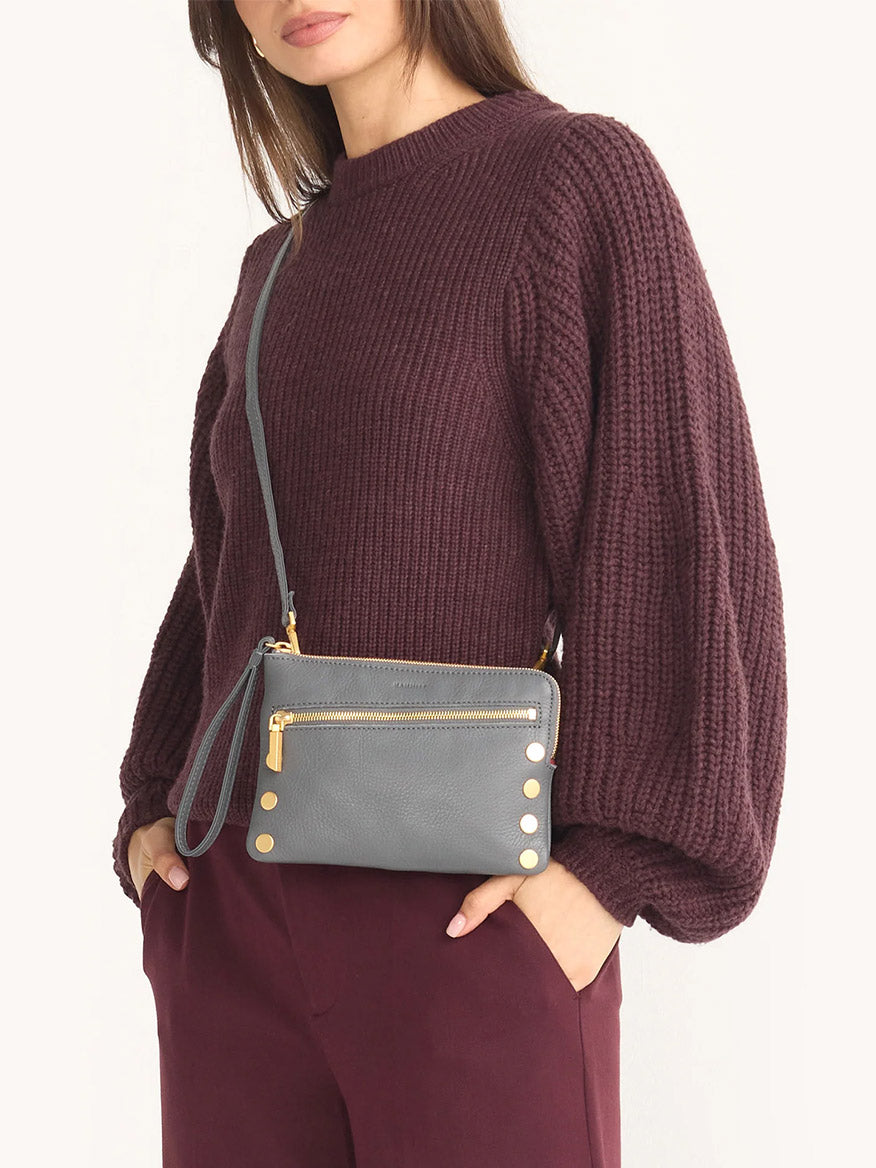 A woman in a textured maroon sweater and pants holds the Hammitt Los Angeles Nash Small Clutch in Sonnet Grey/Brushed Gold, a pebbled leather crossbody with gold zippers and hardware.