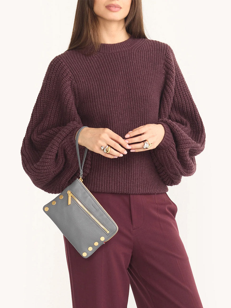A woman in a burgundy knit sweater and pants holds the Hammitt Los Angeles Nash Small Clutch in Sonnet Grey/Brushed Gold, crafted from pebbled leather with gold studs and a zip pocket.