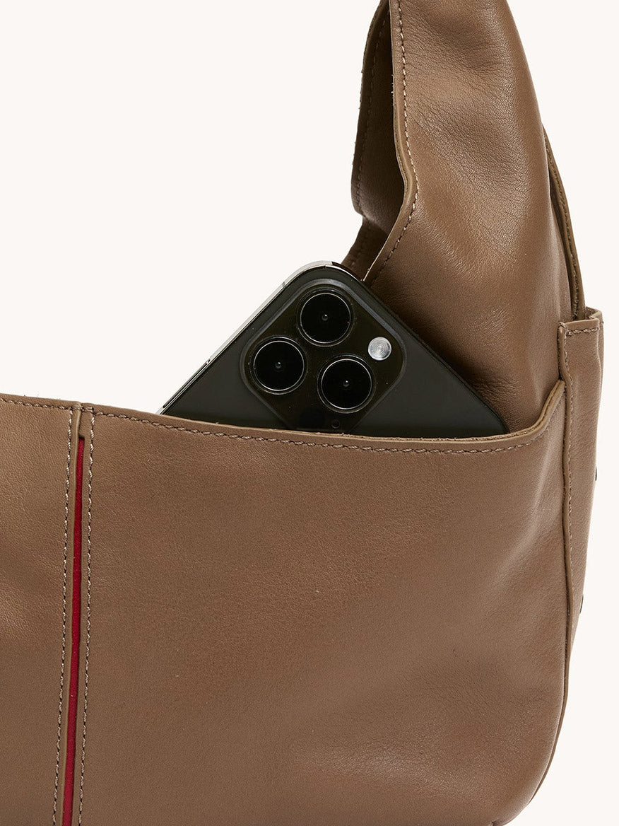 A black smartphone with three lenses peeks out from the outer pocket of the Hammitt Los Angeles Tom Zip Medium in Dune/Silver, a brown leather hobo bag with an adjustable shoulder strap.