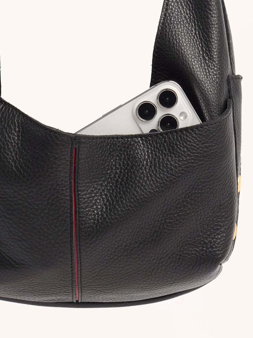 The Hammitt Los Angeles Tom Zip Medium in Black/Brushed Gold & Red Zip is a black pebbled leather hobo bag with an exterior pocket holding a silver smartphone with three rear cameras, featuring an adjustable shoulder strap.