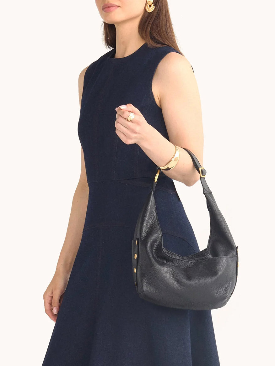A woman in a sleeveless dark blue dress holds the Hammitt Los Angeles Tom Zip Medium handbag in Black/Brushed Gold & Red Zip, featuring pebbled leather, gold accents, and an adjustable shoulder strap against a plain light background.