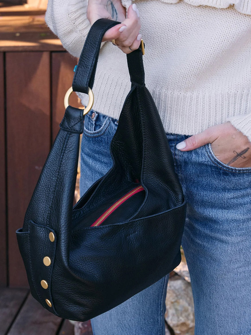 A person in jeans and a cream sweater holds the Hammitt Los Angeles Tom Zip Medium in Black/Brushed Gold & Red Zip, a black pebbled leather hobo bag featuring gold hardware, a red zipper, and an adjustable shoulder strap.