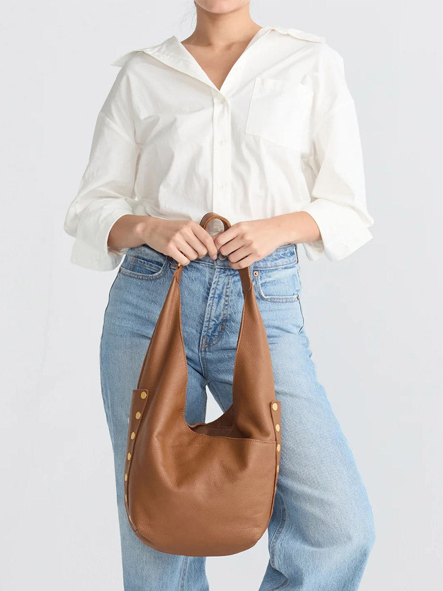 A person in a white blouse and blue jeans holds the Hammitt Los Angeles Tom Zip Large in Mahogany Pebble with brushed gold studs and a red zip, standing against a plain white background.