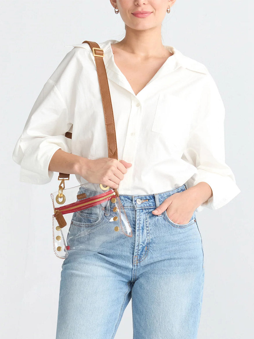 A person in a white button-up shirt and blue jeans holds the Hammitt Los Angeles Tony Small Crossbody Bag in Clear Mahogany Pebble & Red Zip.