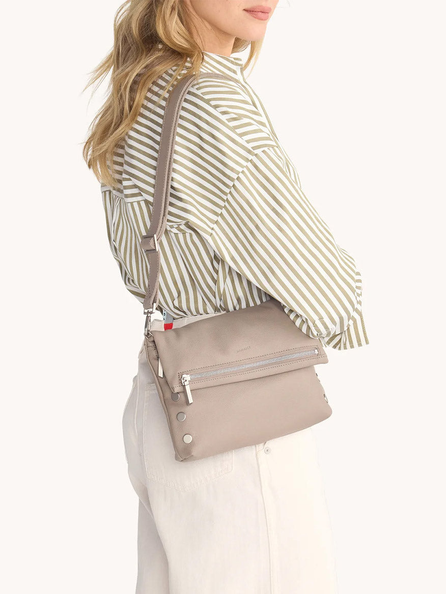 A person wearing a striped shirt and white pants carries the Hammitt Los Angeles VIP Medium Clutch in Pewter Pebble, crafted from pebble leather with silver zippers, snap details, and a removable crossbody strap.