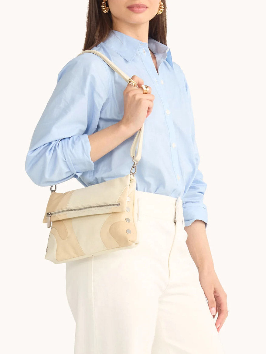 A woman in a light blue button-up shirt and white pants holds the Hammitt Los Angeles VIP Medium Clutch in Coastal Cream/Brushed Silver, standing against a plain background.