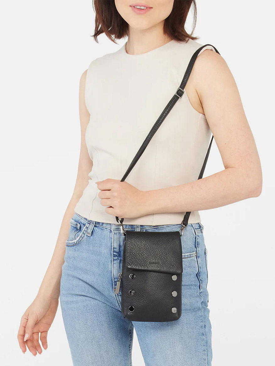 A person in a sleeveless beige top and blue jeans holds the Hammitt Los Angeles VIP Mobile crossbody bag in Black/Gunmetal, featuring pebbled leather, silver studs, and a flap closure.