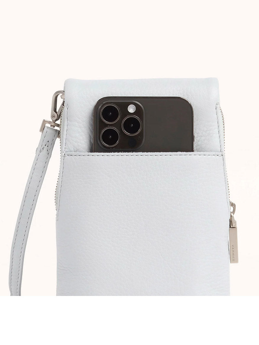 A black smartphone partially tucked into the front pocket of a Hammitt Los Angeles VIP Mobile in Bluebird/Brushed Silver, featuring pastel pebbled leather, a wrist strap, and a visible zipper.